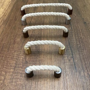 May include: Five rope-style cabinet pulls with metal end caps. The pulls are made of thick, off-white rope and have end caps in various finishes, including silver, gold, and dark brown. The pulls are arranged on a wooden surface.
