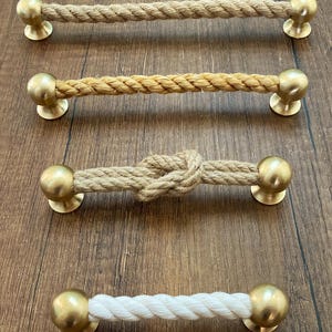 May include: Four drawer pulls featuring a central rope section connected to two spherical brass-toned metal ends. Three pulls have natural tan rope, with one featuring a central knot, while the fourth pull has a white rope section.