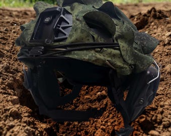 May include: A camouflage helmet cover with a black mount attached to the top. The helmet cover is made of a green and brown fabric with a digital camouflage pattern. The helmet cover is lying on the ground.