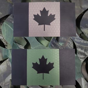 Könnte beinhalten: Zwei kanadische Flaggenaufnäher. Ein Aufnäher zeigt ein schwarzes Ahornblatt auf silbernem Hintergrund mit einem Wabenmuster. Der andere Aufnäher hat ein schwarzes Ahornblatt auf grünem Hintergrund. Beide Aufnäher haben einen schwarzen vertikalen Balken auf der linken Seite.