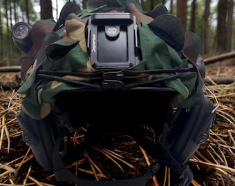Puede incluir: Un casco de camuflaje con un soporte negro para una cámara u otro dispositivo. El casco tiene una correa negra y una luz negra en el lateral. El casco está sobre un lecho de agujas de pino marrones.