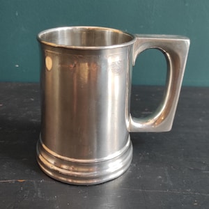 May include: A silver-colored, vintage-style beer mug with a rectangular handle. The mug has a cylindrical body with a slightly flared base and a smooth, reflective surface. The mug is made of metal and sits on a dark wooden surface.