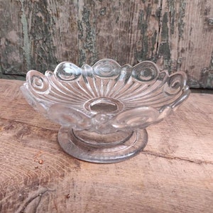 May include: A clear glass pedestal dish with a scalloped edge and a decorative pattern. The dish has a raised centre and a circular base.