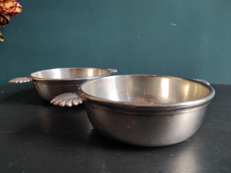 May include: Two silver bowls with handles in the shape of leaves. The bowls are on a black surface.