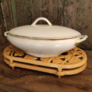 May include: White porcelain serving dish with a gold trim and lid, sitting on a wooden stand with a decorative design.