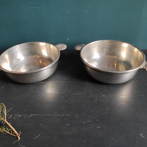 May include: Two silver serving bowls with ornate handles. The bowls are empty and sitting on a dark surface.