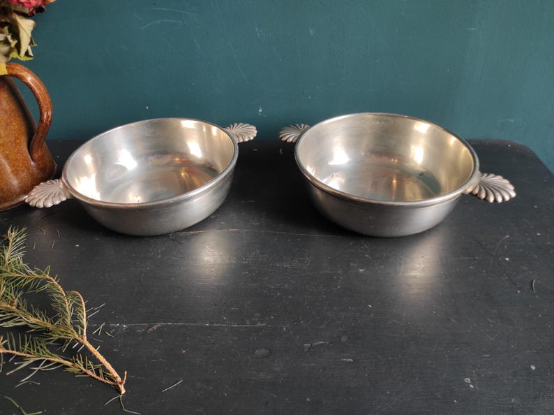 May include: Two silver serving bowls with ornate handles. The bowls are empty and sitting on a dark surface.