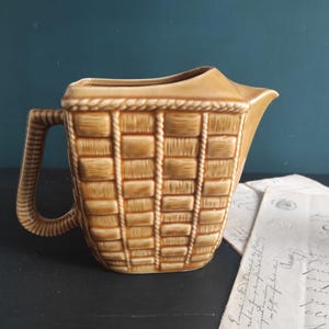 Puede incluir: Una jarra de cerámica marrón con un diseño de cesta tejida. La jarra tiene un asa y un pico.