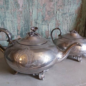 Könnte beinhalten: Ein silbernes Teekannen- und Milchkännchen-Set mit verzierten Blumendesigns und einem erhabenen Wappen. Die Teekanne hat einen Deckel mit einem floralen Knauf und einen Ausguss mit einem gebogenen Griff. Das Milchkännchen hat einen Deckel mit einem floralen Knauf und einem Griff.
