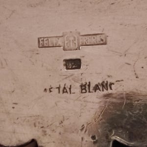 May include: Close-up of a silver-toned metal object with the inscription "Felix et Fr&egrave;res" and "M&eacute;tal Blanc". The object also has the number "1/2" stamped on it.