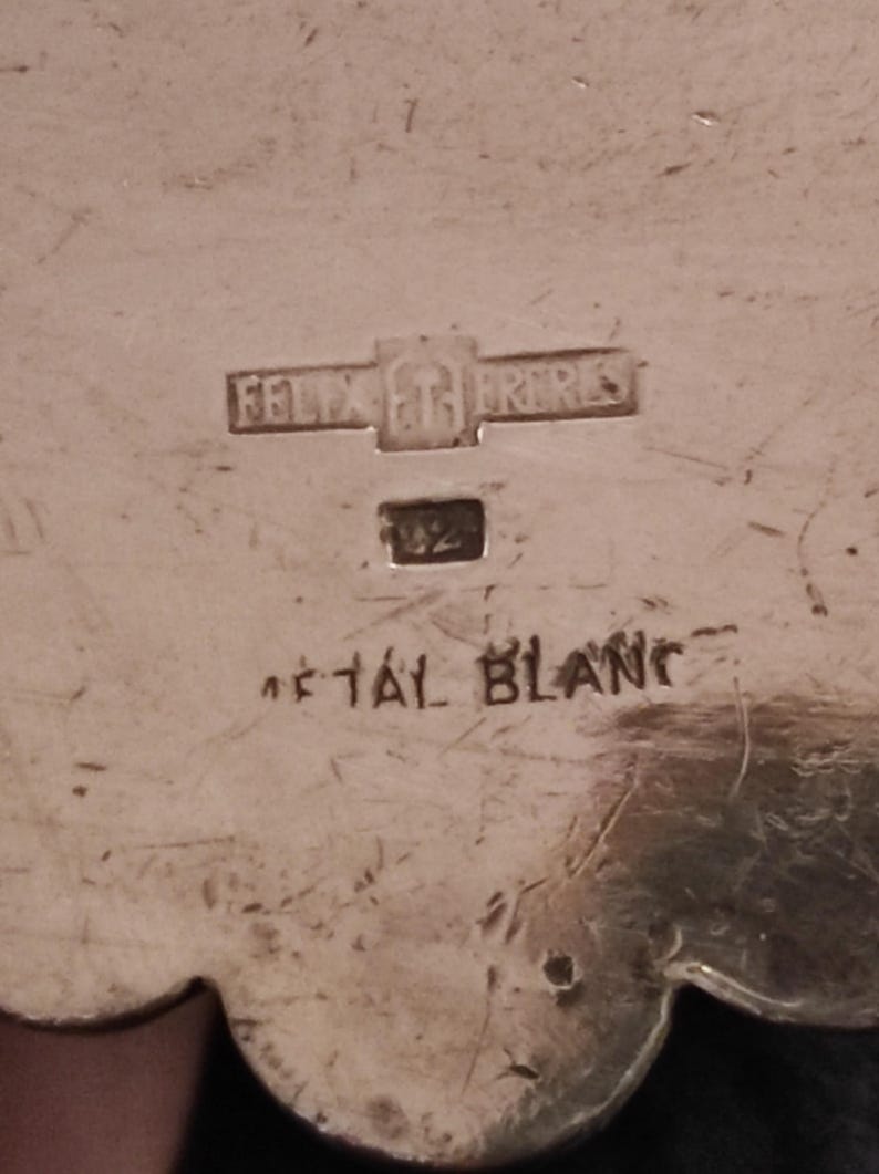 May include: Close-up of a silver-toned metal object with the inscription "Felix et Fr&egrave;res" and "M&eacute;tal Blanc". The object also has the number "1/2" stamped on it.