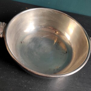 May include: A silver serving bowl with two decorative handles. The bowl is round and has a smooth, shiny surface.