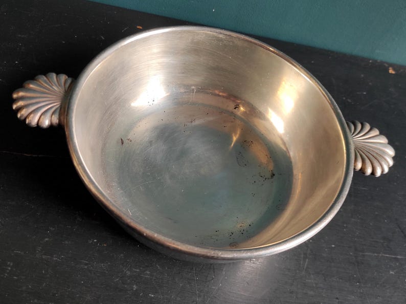 May include: A silver serving bowl with two decorative handles. The bowl is round and has a smooth, shiny surface.