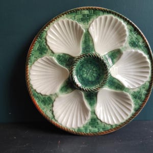 Könnte beinhalten: Runde Austernservierplatte aus Keramik mit sechs weißen, muschelförmigen Fächern und einem zentralen Brunnen. Die Platte hat ein grünes, gewebtes Korbgeflecht-Design mit einem braunen, seilartigen Rand.