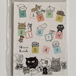 May include: A greeting card with a happy birthday message spelled out with cats sitting on colourful blocks. The card is illustrated with cats in various poses and has the name Shinzi Katoh printed on it.