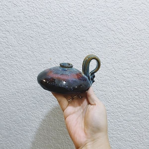 May include: A handmade ceramic oil lamp with a dark blue, red, and brown glaze. The lamp has a rounded body, a small spout, and a curved handle. The lamp is held in a hand.
