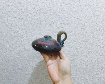 May include: A handmade ceramic oil lamp with a dark blue, red, and brown glaze. The lamp has a rounded body, a small spout, and a curved handle. The lamp is held in a hand.