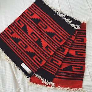 May include: A red and black woven rug with a geometric pattern. The rug has fringe on all sides.