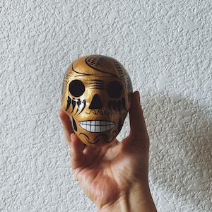 May include: A gold skull with black painted details, including eyes, nose, and mouth. The skull is decorated with intricate patterns and designs.