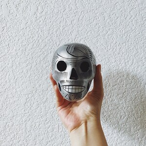 May include: A silver skull with black painted details, including eyes, nose, and mouth. The skull is decorated with a floral pattern.