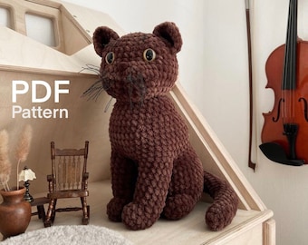 May include: A brown crocheted cat amigurumi toy sitting on a wooden surface. The cat has large brown eyes and a long tail. The text "PDF Pattern" is visible in the image.