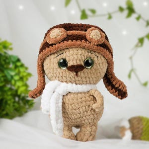 Puede incluir: Un oso de peluche de crochet marrón que lleva un gorro de aviador marrón con orejeras y una bufanda blanca. El oso tiene grandes ojos negros y una nariz marrón.