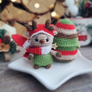 Puede incluir: Una figura de reno de ganchillo con gorro y bufanda rojos, y un atuendo verde, está de pie junto a un árbol de Navidad de ganchillo. El reno tiene cuernos marrones y un botón en forma de estrella. Los artículos están sobre un plato blanco.