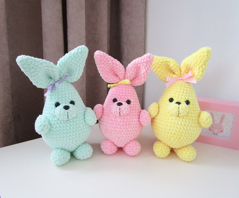 Op de afbeelding: Drie gehaakte konijnenknuffels in verschillende kleuren: lichtblauw, roze en geel. Elk konijn heeft een lang, slap oor en een kleine, zwarte neus.