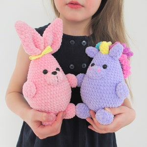 May include: A child holds two crocheted stuffed animals. The pink bunny has yellow ears and a yellow bow. The purple unicorn has a rainbow horn and a pink mane.