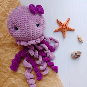 Puede incluir: Un juguete de medusa de crochet morado con un lazo rosa en la parte superior. La medusa tiene largos tentáculos rizados.