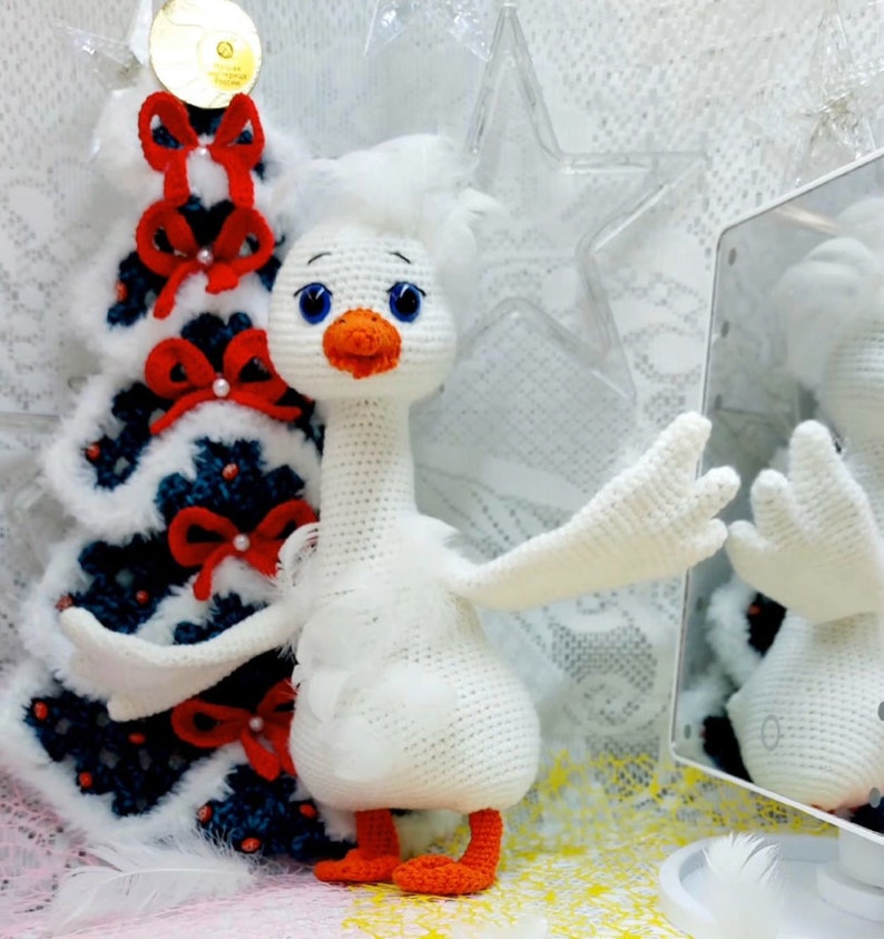 May include: A white crocheted goose toy with orange feet and blue eyes. The goose is standing next to a white and blue Christmas tree with red bows. The goose is looking at its reflection in a mirror.