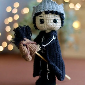 May include: A crocheted doll wearing a grey crown and a black cape, holding a brown and black crocheted rocking horse.