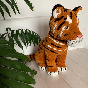 Puede incluir: Un juguete de peluche de tigre hecho a mano en tonos naranja, marr&oacute;n, negro y blanco. El tigre tiene rayas negras, detalles blancos y una nariz rosa. El juguete est&aacute; sobre una superficie blanca con follaje verde en el fondo.