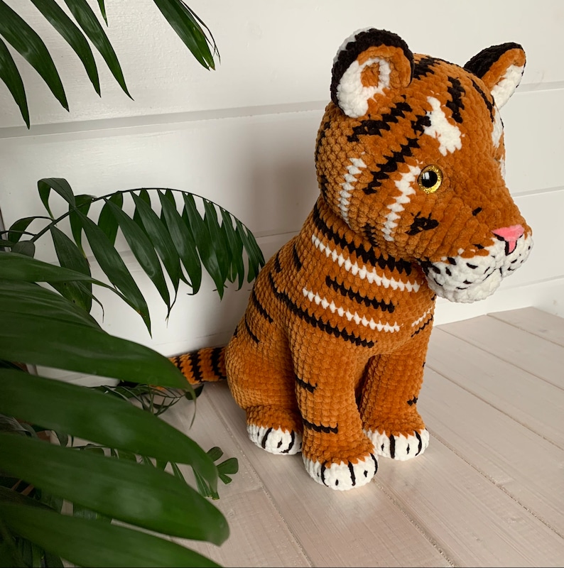 Puede incluir: Un juguete de peluche de tigre hecho a mano en tonos naranja, marr&oacute;n, negro y blanco. El tigre tiene rayas negras, detalles blancos y una nariz rosa. El juguete est&aacute; sobre una superficie blanca con follaje verde en el fondo.