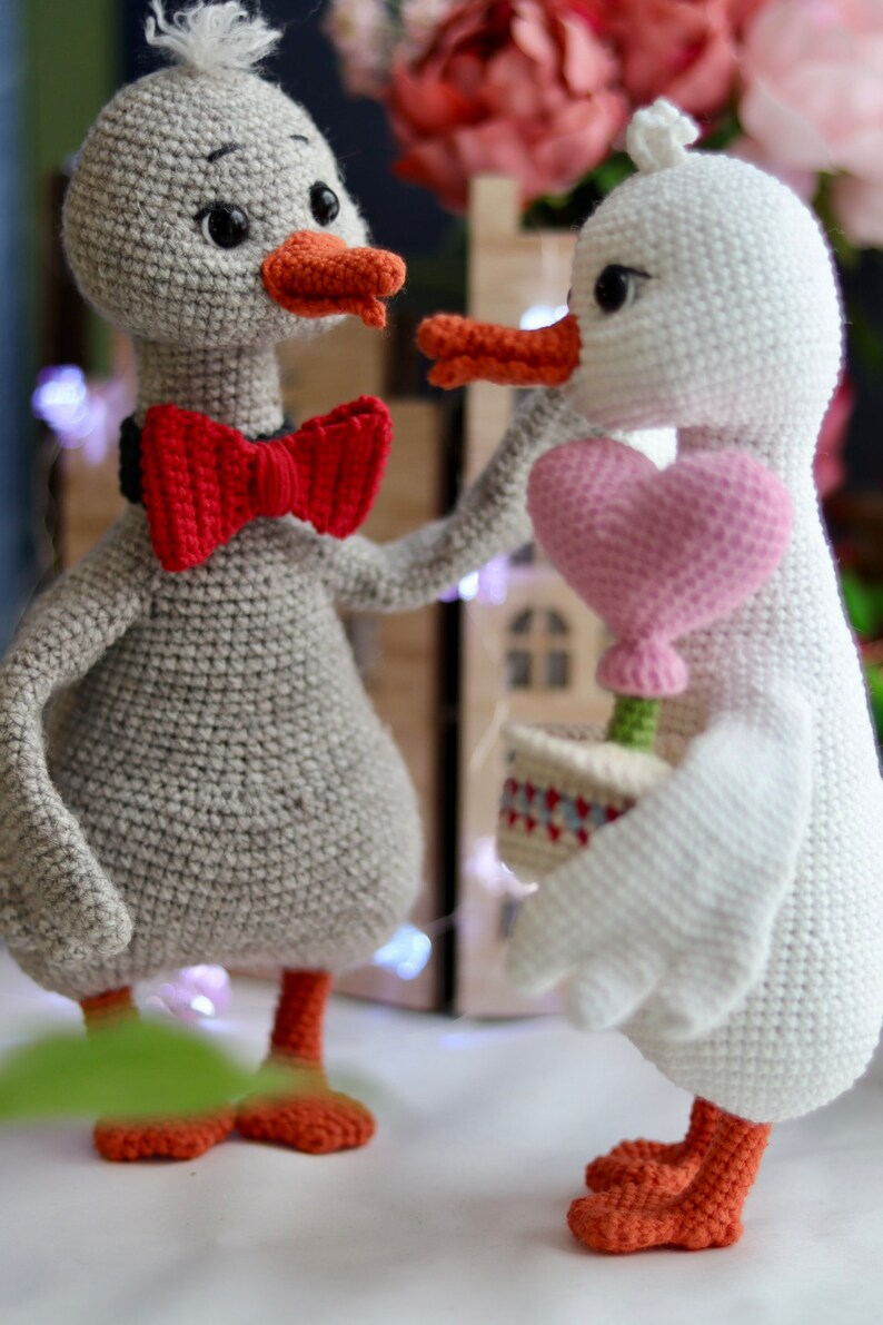 May include: Two crocheted duck plushies, one gray with a red bow tie and the other white holding a pink heart. The ducks are standing on a white surface.