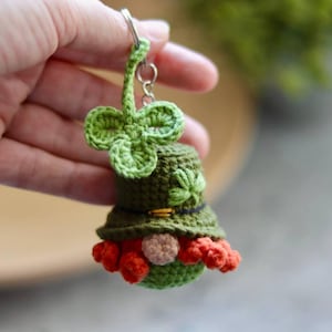 Puede incluir: Un llavero de duende verde tejido a crochet con un amuleto de trébol, un sombrero verde y una barba roja.