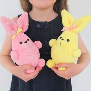 Op de afbeelding: Een kind houdt twee gehaakte konijnenknuffels vast. E&eacute;n konijn is roze met een gele strik en de andere is geel met een roze strik.
