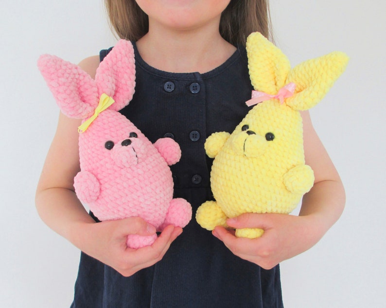 Op de afbeelding: Een kind houdt twee gehaakte konijnenknuffels vast. E&eacute;n konijn is roze met een gele strik en de andere is geel met een roze strik.