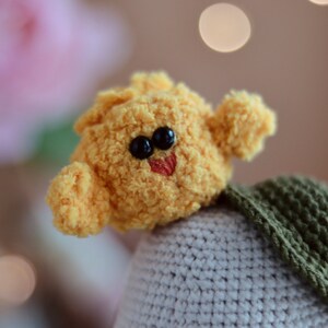 May include: A yellow crocheted plush toy with black eyes and a red mouth, sitting on a gray and green crocheted base.