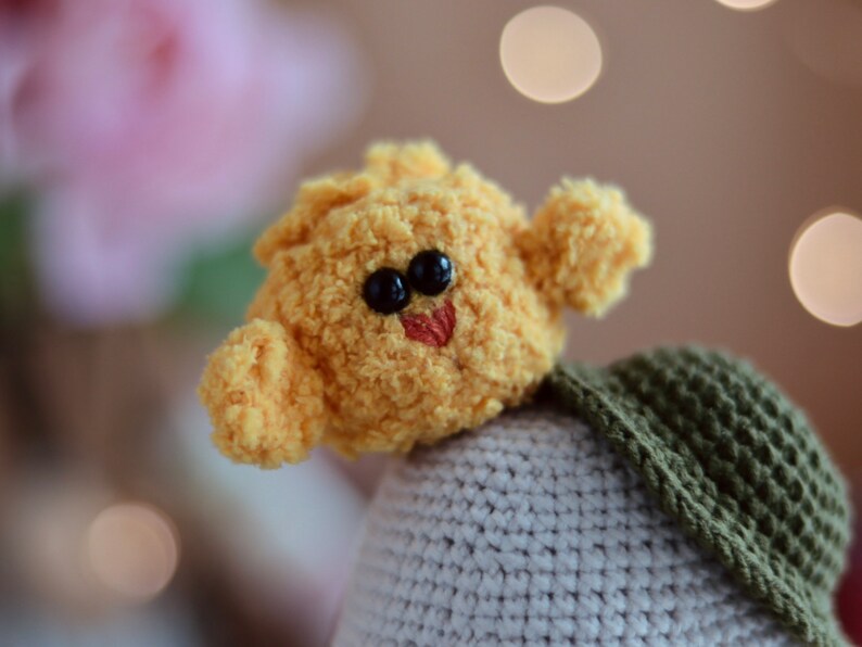 May include: A yellow crocheted plush toy with black eyes and a red mouth, sitting on a gray and green crocheted base.