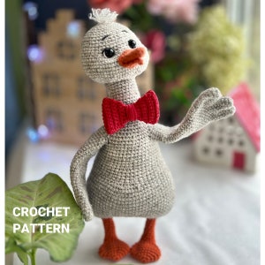 May include: Crochet pattern for a gray amigurumi goose wearing a red bow tie. The goose has a white tuft of feathers on its head and orange feet.