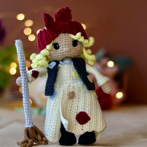 May include: A crocheted doll with blonde hair, a red hat, and a white dress holding a broom. The doll is wearing a black waistcoat with white polka dots.
