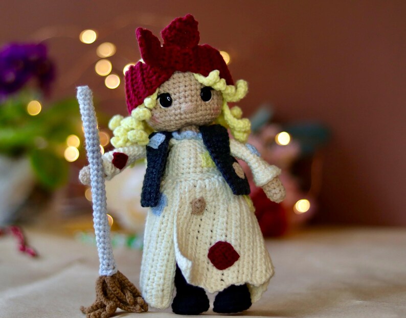 May include: A crocheted doll with blonde hair, a red hat, and a white dress holding a broom. The doll is wearing a black vest with white polka dots.