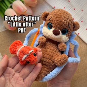 Op de afbeelding: Een bruine gehaakte otterknuffel met grote blauwe ogen en een crèmekleurige snuit, zittend in een lichtblauw gehaakt tasje. Een oranje en witte gehaakte vis zit vast met een koordje. Tekst: Crochet Pattern "Little otter" PDF.