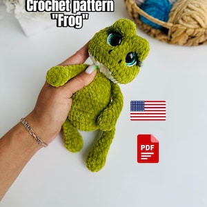 May include: A green crocheted frog with large eyes and eyelashes is held in a hand. The text "Crochet pattern "Frog"" is at the top. An American flag and a PDF icon are also visible.