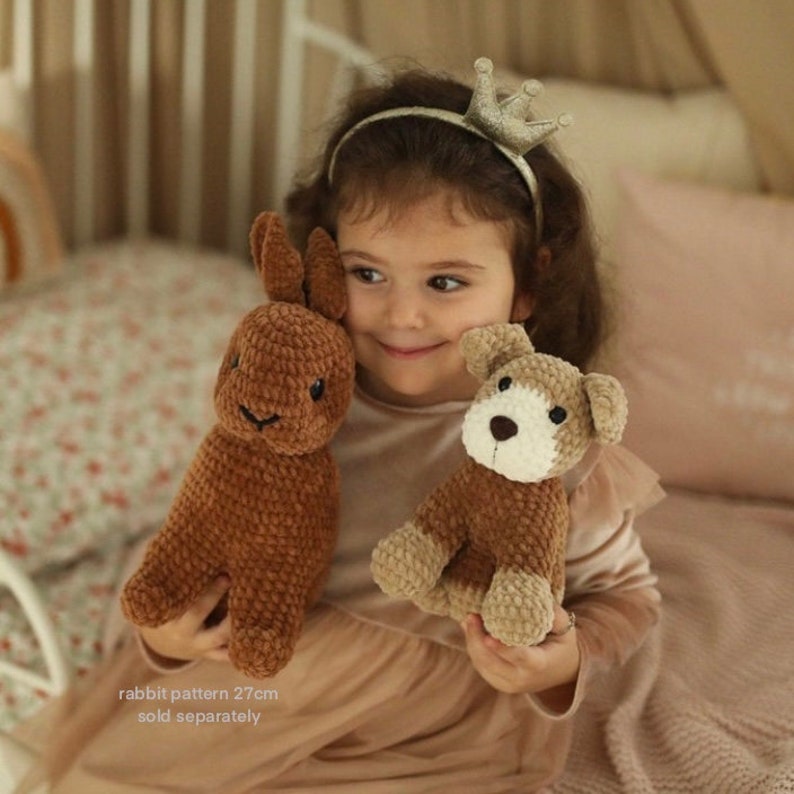 May include: A young child wearing a crown headband holds a brown crocheted rabbit and a brown crocheted dog. The rabbit is 27 cm tall and is sold separately.