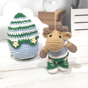 May include: A crocheted moose wearing a blue and green striped shirt and green shoes. The moose is standing next to a crocheted egg with green and white stripes.