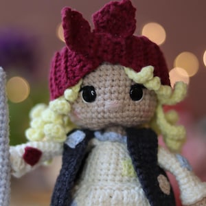 May include: A crocheted doll with blonde hair, wearing a red and white crocheted hat and a brown vest. The doll is holding a gray crocheted stick.