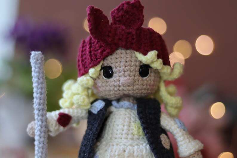 May include: A crocheted doll with blonde hair, wearing a red and white crocheted hat and a brown vest. The doll is holding a gray crocheted stick.