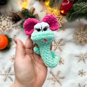 Può includere: Un giocattolo a forma di serpente verde all'uncinetto con orecchie rosa e macchie gialle. Il serpente ha un viso bianco con ciglia nere e occhi marroni. Il giocattolo è tenuto in una mano.
